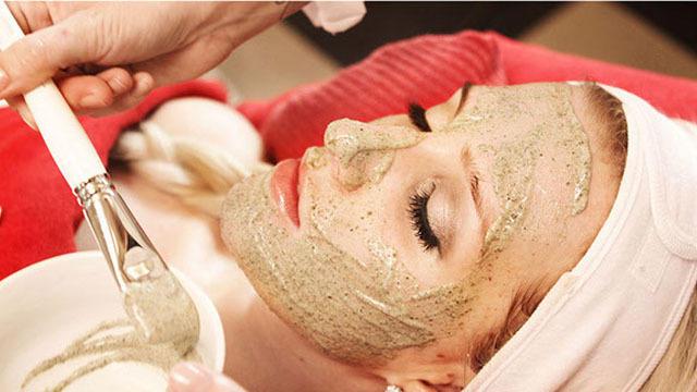 Kvinne får ansiktsmaske på spa, med grønn leiremaske og pensel i hånden.