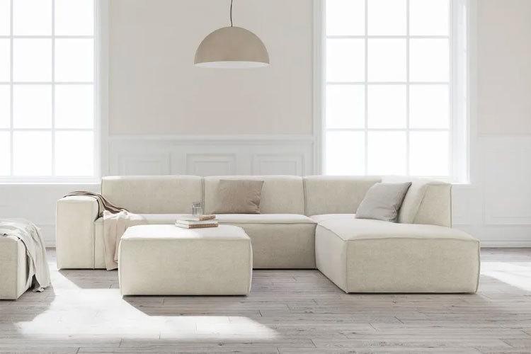 Lys beige hjørnesofa med puter og et kvadratisk bord i et rom med store vinduer og lys innredning.