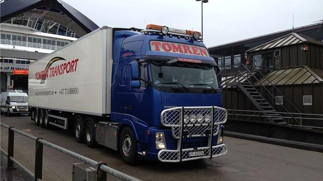 Blå lastebil med påbygg for transport, parkert ved en bygning med moderne arkitektur.