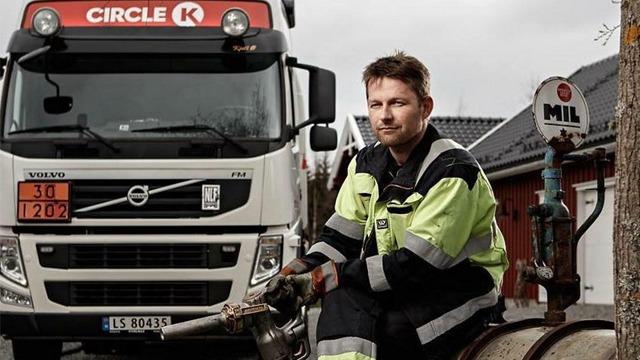En mann i arbeidsklær sitter på en bensinstasjon med en lastebil i bakgrunnen.