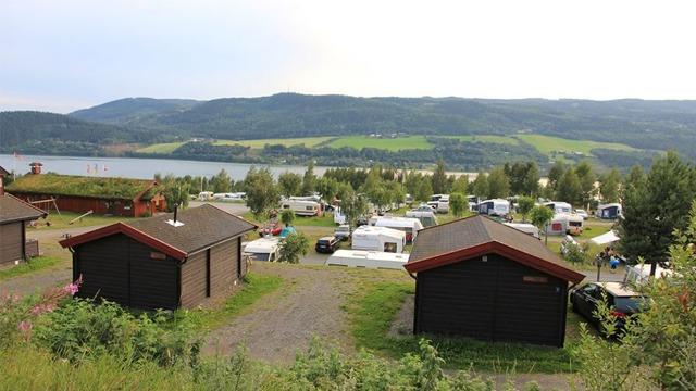 Utsikt over campingplass med hytter og campingvogner ved innsjøen, omkranset av grønne åser.