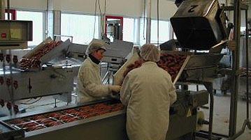 To personer i hvite arbeidsklær jobber med pølsefabrikkmaskiner i en produksjonslinje.