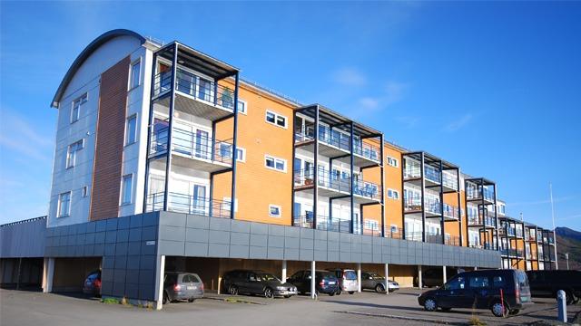 Moderne leilighetsbygg med balkonger og parkeringsplasser under, beliggende i solfylt område.