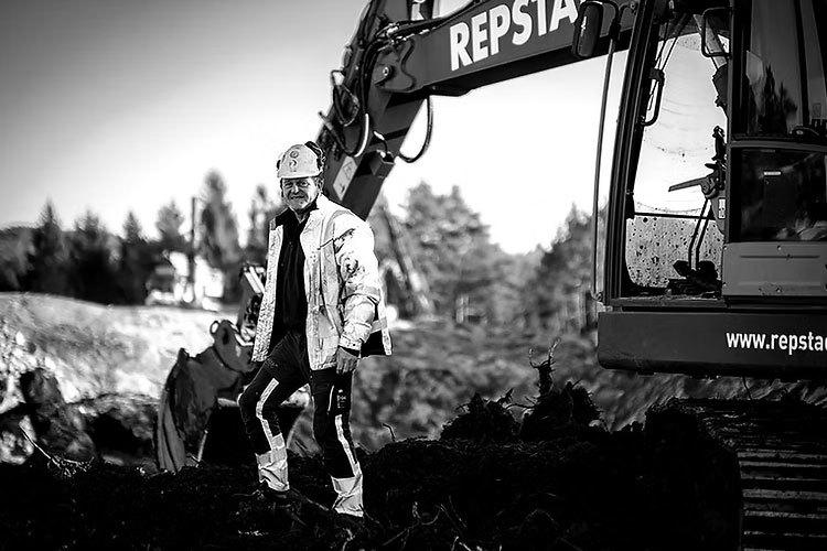 En mann står ved en gravemaskin med "REPSTA" logo, iført arbeidsklær og hjelm, i et byggeområde.