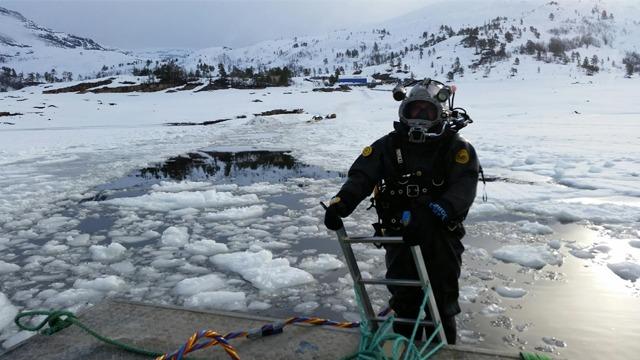 Dykker i dykkerdrakt står på en plattform ved isdekket vann, med isflak rundt seg.