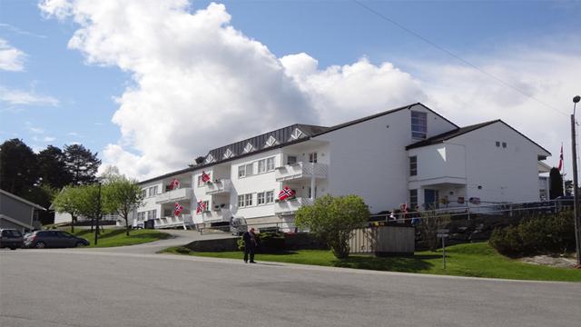 Moderne bygning med hvit fasade og balkonger, omgitt av grønt landskap og blå himmel med skyer.