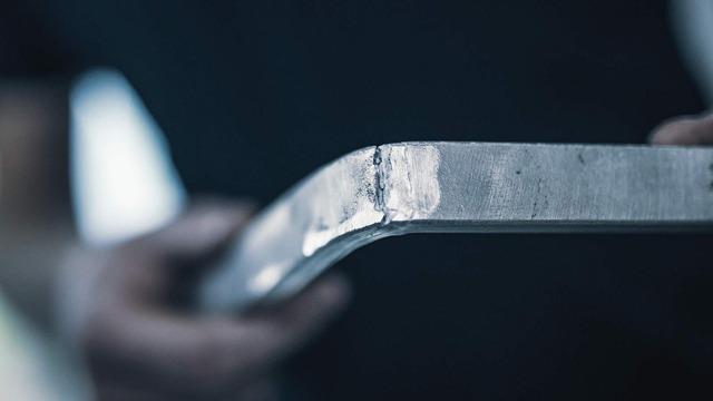En person holder en metallstang med en buet ende, fokusert på sveisesømmen.