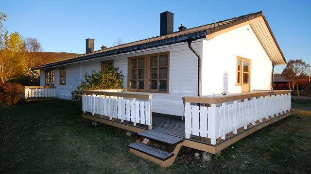 Et hvitt hus med veranda og trapp, omringet av grønt gress og trær i bakgrunnen. Sollys skinner på fasaden.