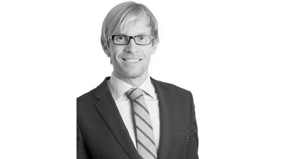 En smilende mann med briller iført dress og slips, står foran en lys bakgrunn.