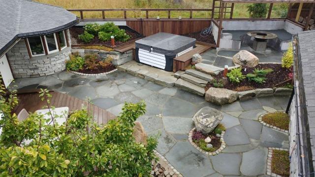 Hage med steinlagt terrasse, jacuzzi, planter og en sitteplass med steinmur.