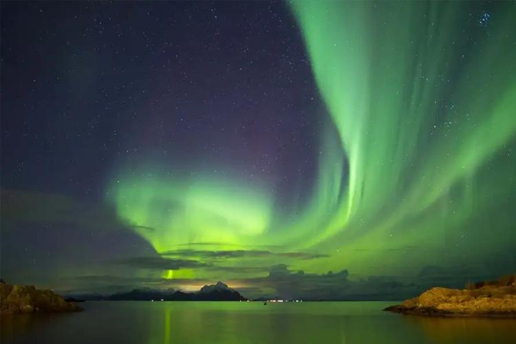 Nordlys som lyser opp nattehimmelen over en rolig sjø med fjell i bakgrunnen.