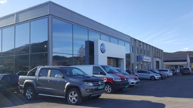 Bilforhandler med flere biler parkert foran bygningen og store vinduer som reflekterer himmelen.