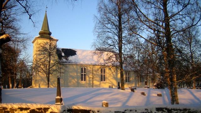 En vakker, snødekt kirke med spire, omringet av trær i sollys. Gravsteiner synlige i forgrunnen.