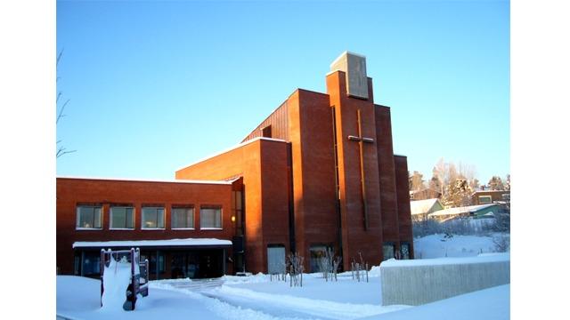 Moderne kirke med rød murstein og snødekte omgivelser under en klar blå himmel.