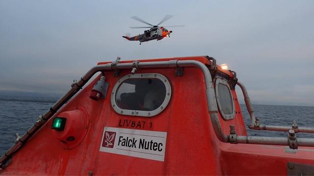 Rød redningsbåt med Falck Nutec-logo og helikopter i luften over havet under en grå himmel.