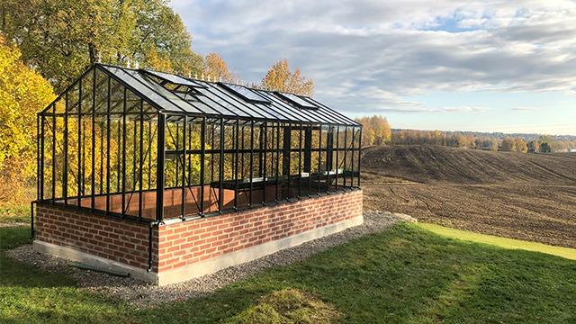 Et moderne drivhus i glass og murstein, plassert på en grønn bakke med høstfarger i bakgrunnen.