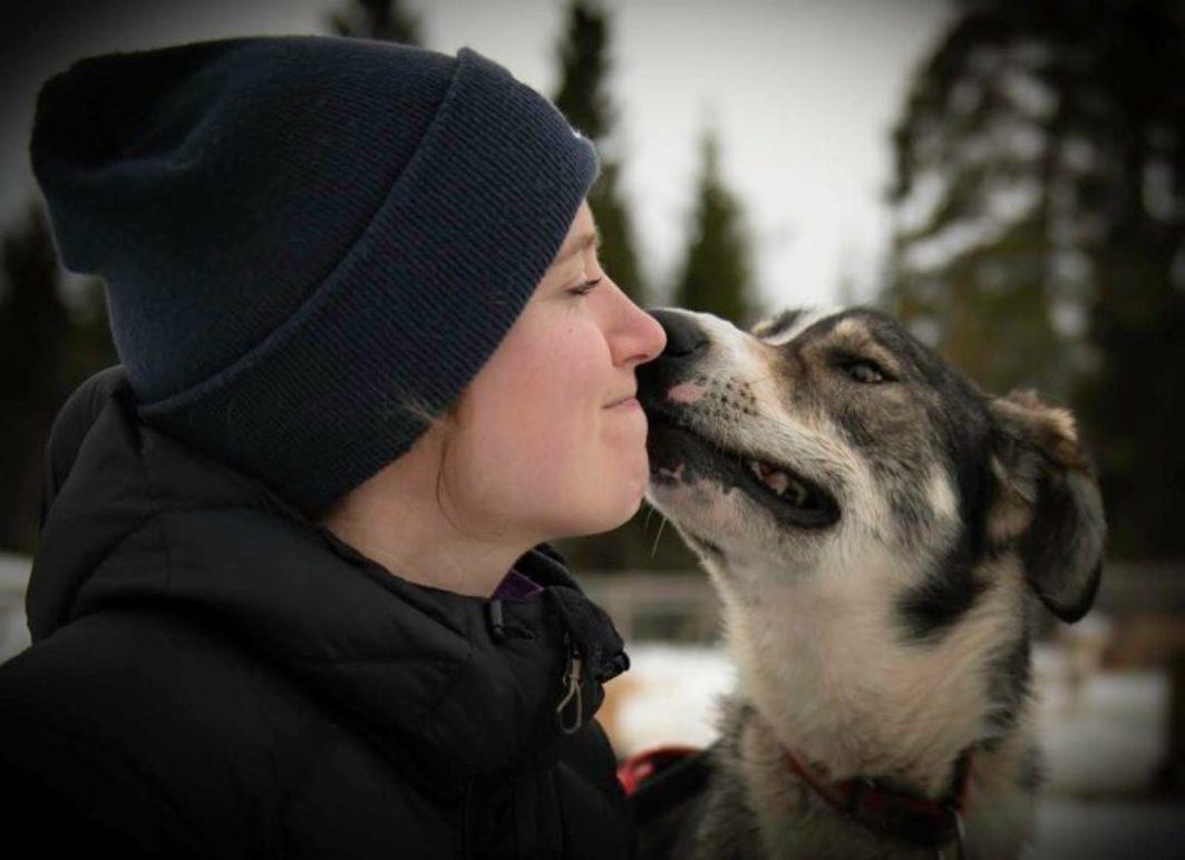 En kvinne i vinterklær smiler mens en hund kysser henne på kinnet i en snødekt skog.