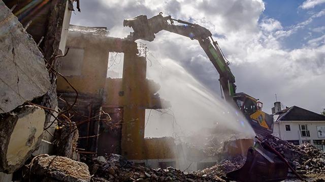 Rivemaskin sprayer vann på en bygning under riving, med grå skyer i bakgrunnen.