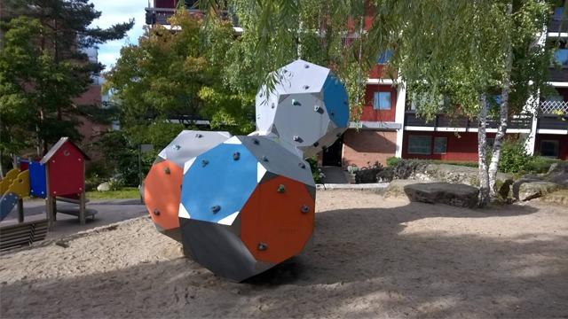 Lekemodul i sandkasse med geometriske former i farger som blå, oransje og grå, omgitt av trær og bygninger.