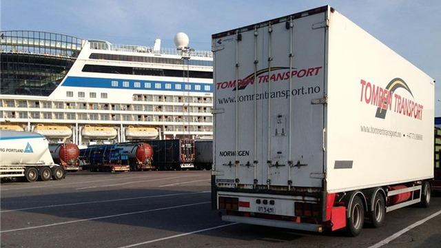 Lastebil fra Tomars Transport parkert ved en cruiseskip i havnen, med flere lastebiler i bakgrunnen.