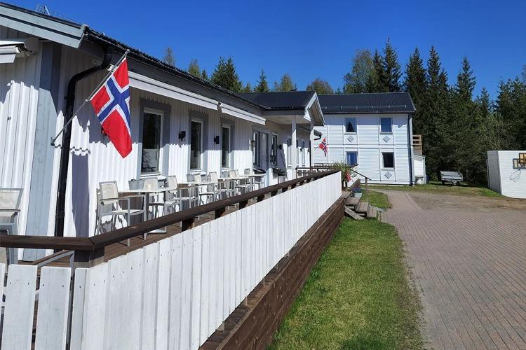 En bygning med norsk flagg, uteplass med stoler og bord, omgitt av trær og blå himmel.