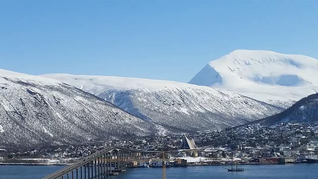 Vinterlandskap med snødekte fjell, en bro og en liten by ved vannet under klar blå himmel.