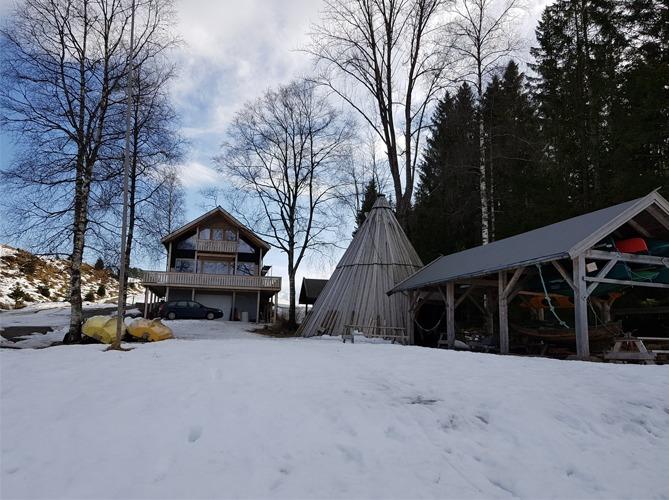 Hus med balkong, lavvo og uthus i snødekt landskap med trær i bakgrunnen.
