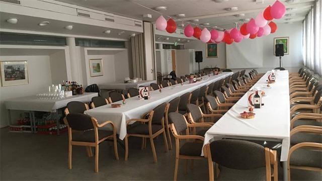 Lokal med langbord dekket til fest, pyntet med rosa ballonger og stoler i en lys atmosfære.