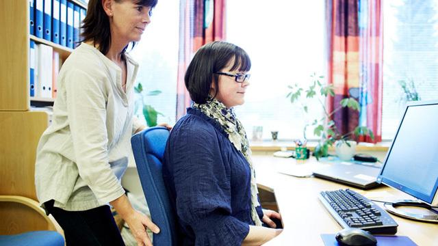 To kvinner i et kontor, hvor en hjelper den andre med å justere stolen foran en datamaskin.