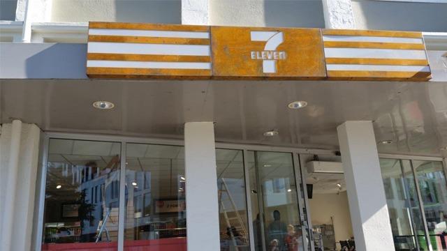 7-Eleven butikk med stor, oransje logo og glassfasade. Solfylt dag med mennesker inne.