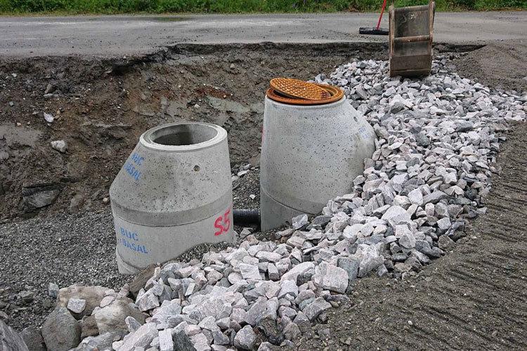 To store betongrør og grus ved en vei, med en åpen grøft og en jernlokk på toppen av røret.