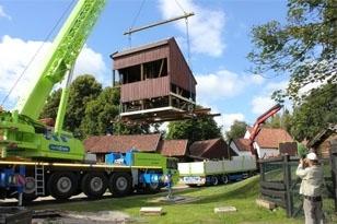 En stor kran løfter et hus mens det transporteres på en lastebil, med trær og bygninger i bakgrunnen.