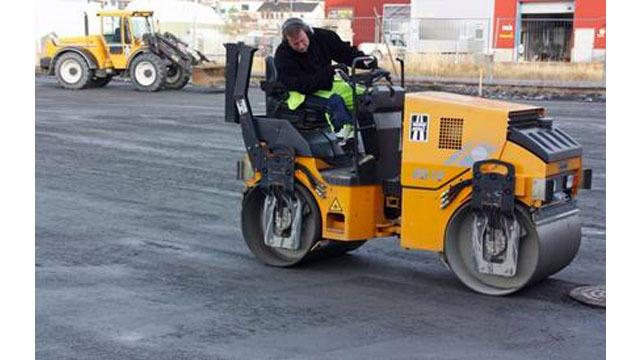 En mann kjører en asfaltvalser på en byggeplass, med en traktor i bakgrunnen.