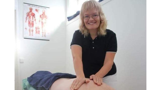 En smilende kvinne gir massasje til en pasient på en behandlingsbenk i et rom med anatomiske plakater.