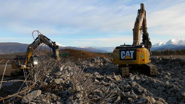 To store og kraftige gravemaskiner jobber på en steinete byggeplass med fjell i bakgrunnen.