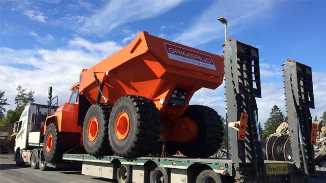 En stor oransje dumper på en lastebiltrailer under en blå himmel med skyer.