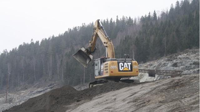 Gravemaskin i arbeid på en skråning med skog i bakgrunnen. Maskinen har en CAT-logo på siden.