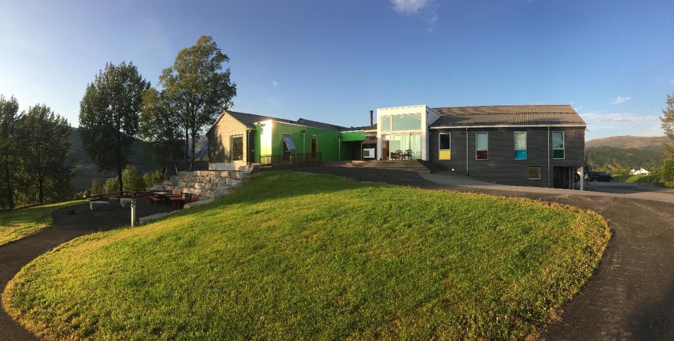 Moderne hus med grønn fasade, omgitt av grønt gress og trær, under en klar blå himmel.