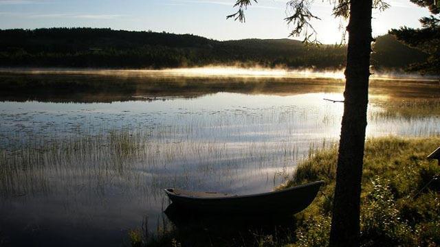 En stille innsjø med tåke, en båt ved bredden og trær som rammer inn landskapet i morgenlyset.