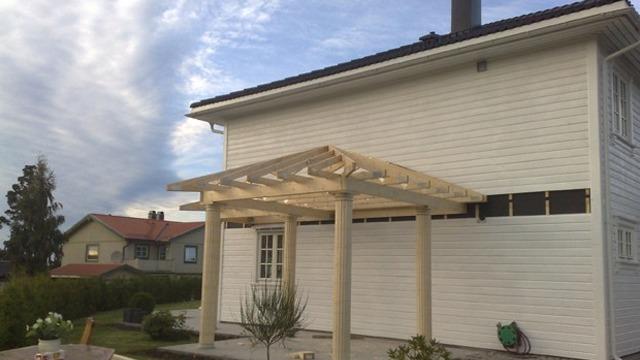 Hvit enebolig med pergola på terrassen, omgitt av grønt landskap og blå himmel.