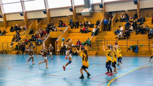 Barn som spiller håndball i en idrettshall, med tilskuere som heier fra tribunen.