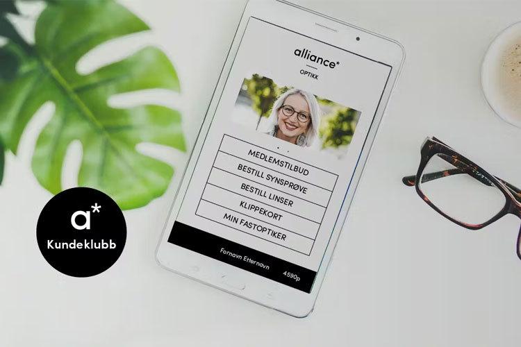Bilde av en nettbrett som viser en app med menyvalg for kundeklubb, med briller og en plante i bakgrunnen.