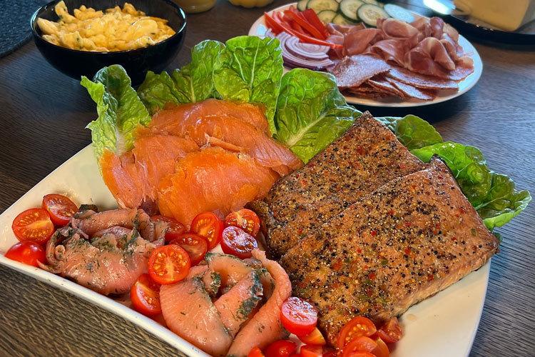 Et fat med røkt laks, stekt laks, cherrytomater og salat, servert med skiver av skinke og grønnsaker i bakgrunnen.