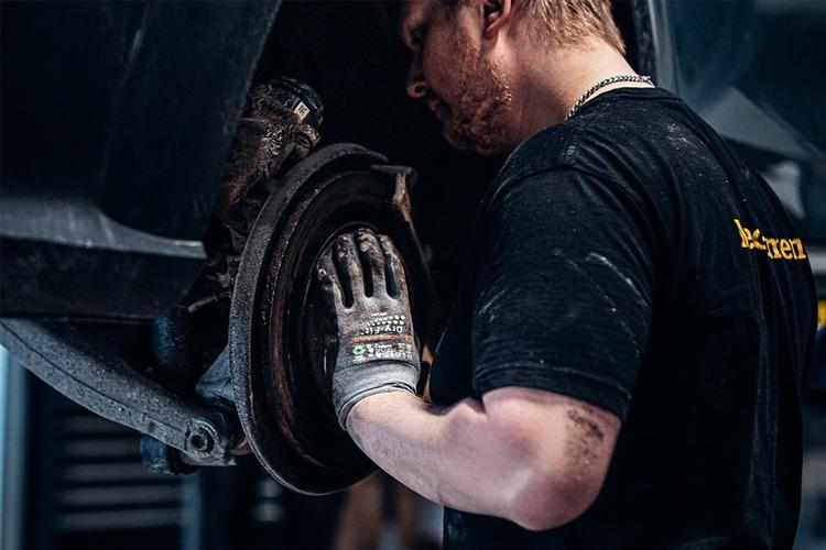En mekaniker jobber med bremsene på en bil i et verksted, iført arbeidshansker og svart t-skjorte.