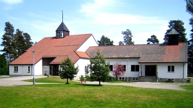 En hvit kirke med rød tak og en tilstøtende bygning, omgitt av trær og grønt gress under en blå himmel.