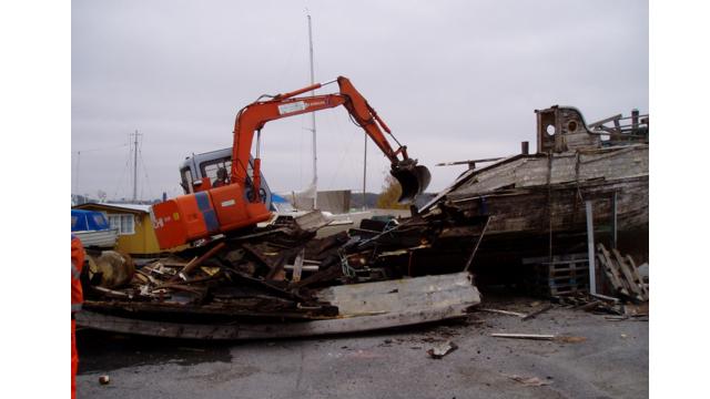 En gravemaskin fjerner deler av et gammelt skip på en havn, med grå himmel i bakgrunnen.