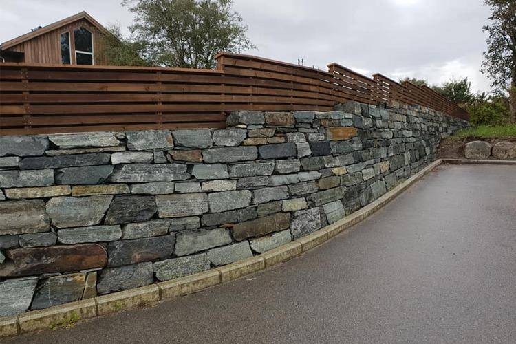 En steinmur langs en vei med trehegn og et hus i bakgrunnen under en skyet himmel.