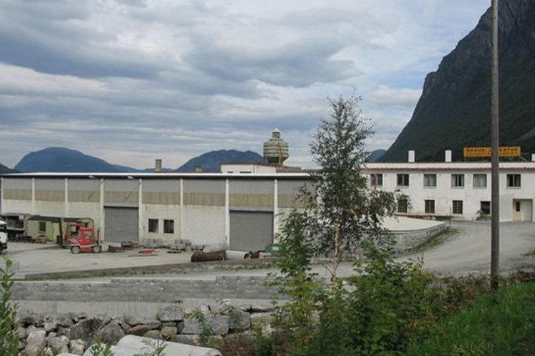 En stor bygning med flere vinduer og en grå bakgård, omgitt av fjell og trær i bakgrunnen.
