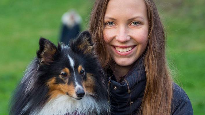 En kvinne smiler mens hun holder en colliehund i armene, med grønt gress i bakgrunnen.