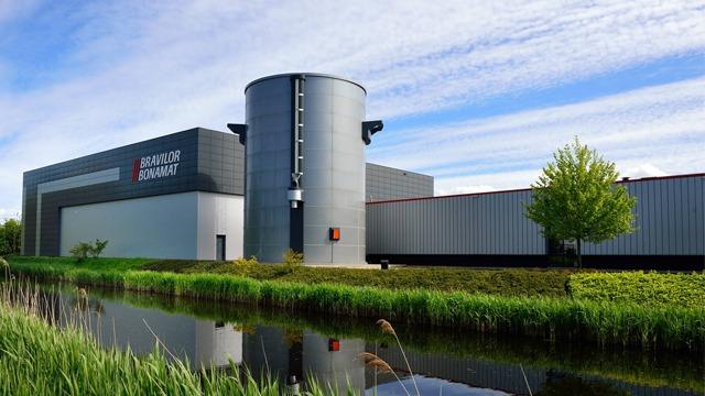 Moderne industribygg med silotårn ved en grønn elv under en blå himmel.